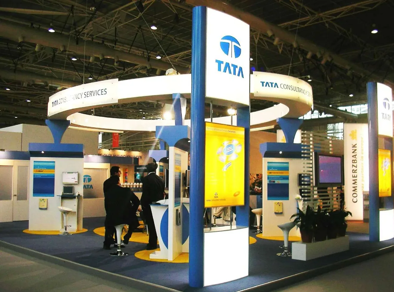 A circular TCS exhibition pavilion with a suspended white fascia ring carried on tapered blue columns, backlit graphic panels and yellow floor discs