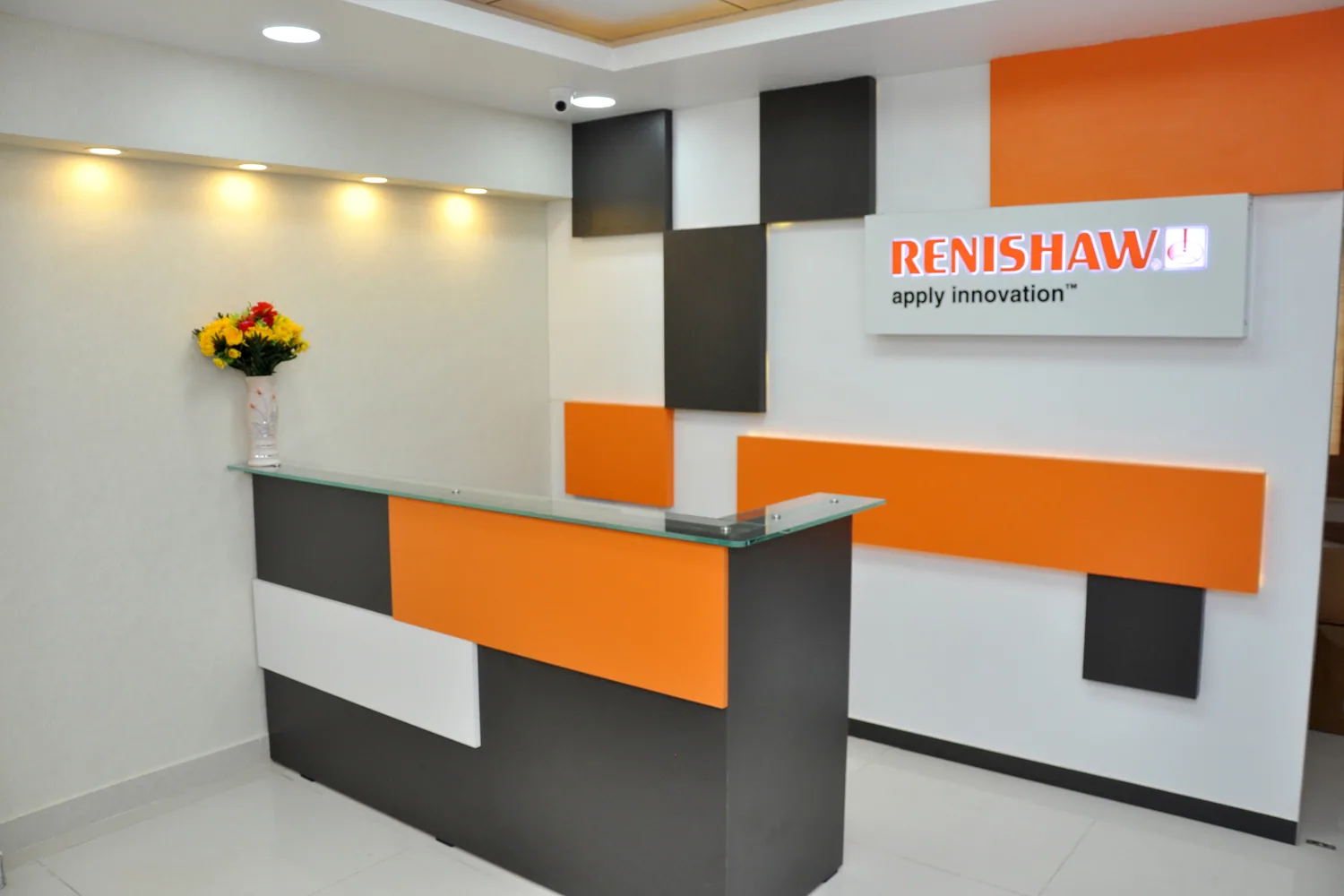A workplace reception with a charcoal and orange laminate desk in front of a composition wall of offset orange, white and grey panels beneath an illuminated logo